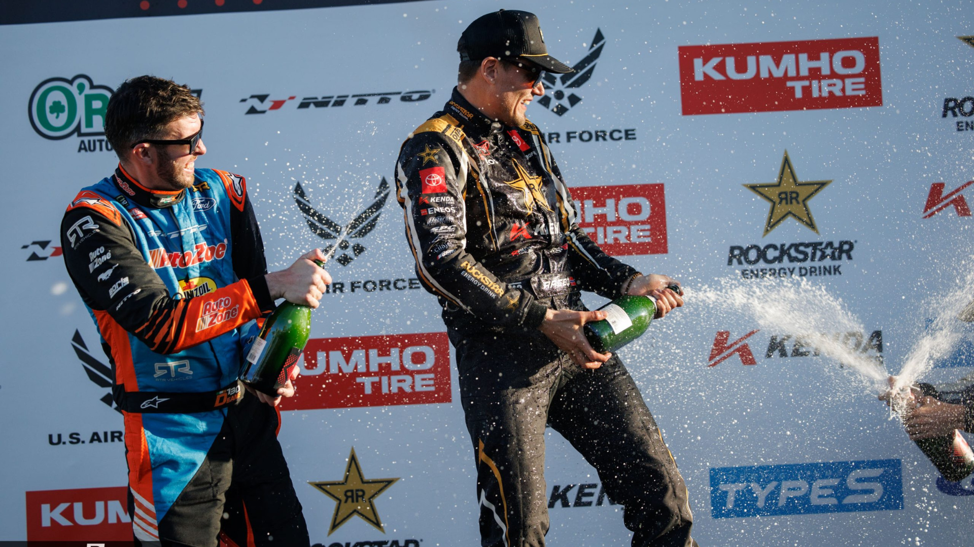 Drivers celebrating with champagne in front of a logo-filled backdrop